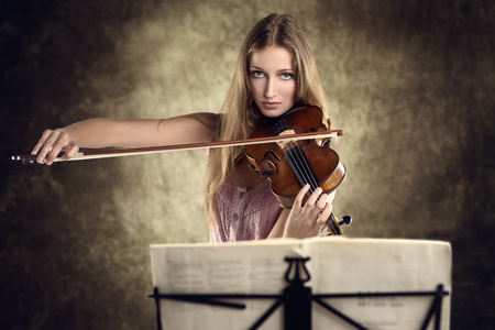 Pretty young female violinist in a stylish pink outfit standing playing the violin as she gives a classical recital at the academy or practices for a performanceの写真素材