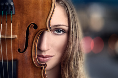 Close up Pretty Young Woman Peeking Behind Violin Instrument in front of lights in the evening or nightの写真素材