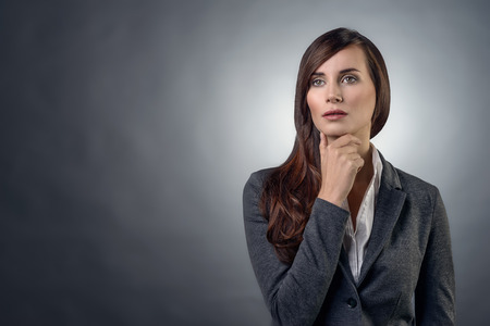 Thoughtful serious businesswoman with an inscrutable expression staring straight ahead with her hand to her chin, over grey with copy spaceの写真素材