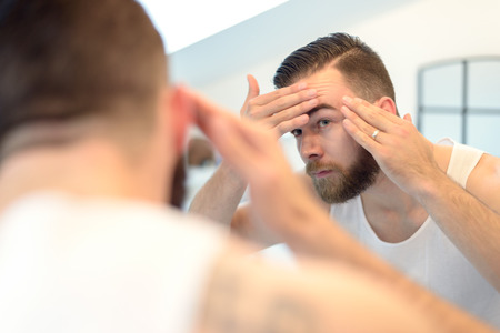 Bearded man checking his complexion in the bathroom mirror, looking concerned, rear view over the shoulder.の写真素材