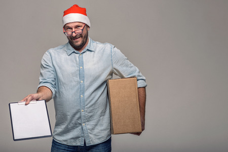 Cheerful attractive young man in a Santa Hat carrying or delivering a large brown cardboard box for Christmas, over grey with copy spaceの写真素材