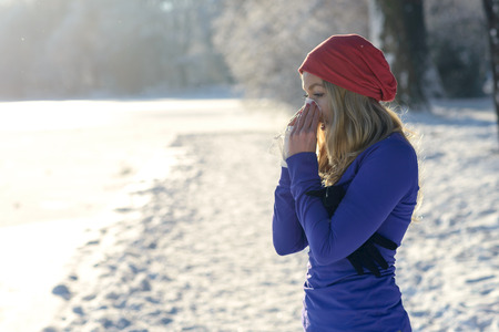 Young woman suffering from a winter cold standing outdoors in snowy cold countryside blowing her nose on a handkerchief or tissue, with copy space over pristine white snowの写真素材