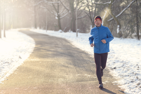 Man out in winter weather for his daily run jogging on a paved road in a snowy landscape in a fitness and healthy lifestyle concept, close up full bodyの写真素材