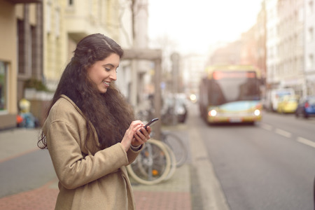 Texting brunette woman reads a message on her mobile phone and smiles while being unaware of approaching busの写真素材
