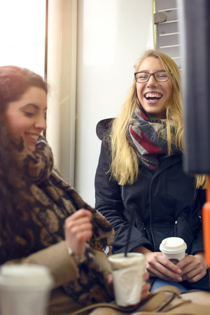 Two seated blond and brunette female friends wearing coats and holding coffee share a humorous momentの写真素材