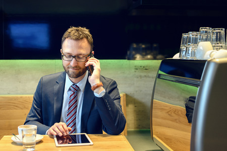 Handsome businessman with striped tie sits at table with coffee making phone call and using his tabletの写真素材