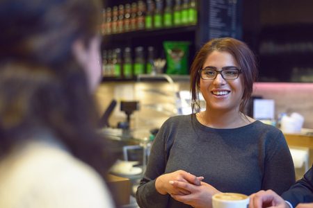 Young waitress serving a cup of coffee to a pretty young female customer in a restaurant or pub placing the cup on the table with a smileの写真素材