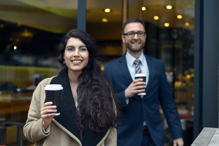 Two business colleagues purchasing takeaway coffee leaving a coffee house or bistro with the disposable cups in their hands as they smile at the cameraの写真素材