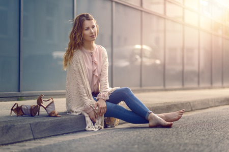 Serious pretty trendy young woman sitting relaxing on a sidewalk on an urban street in her high heels and fashionable outfitの写真素材