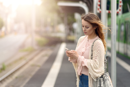 Attractive stylish young woman waiting alone on a platform at a small urban station checking her mobile phone for messagesの写真素材