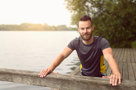 Young man doing stretching exercises on a wooden deck overlooking a lake as he smiles at the camera in a healthy lifestyle conceptの写真素材