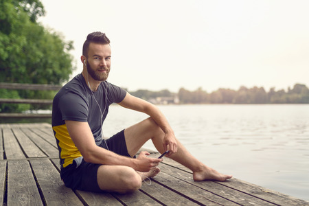 Attractive barefoot bearded young man relaxing on a wooden deck overlooking a calm lake with his mobile phone in his hand looking at the camera with a smileの写真素材
