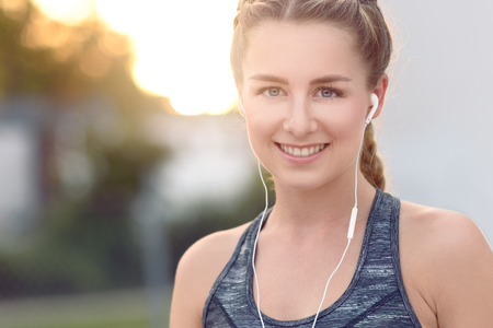Attractive young woman with a lovely smile listening to music on ear plugs on a summer evening with the glow of the setting sun behind herの写真素材