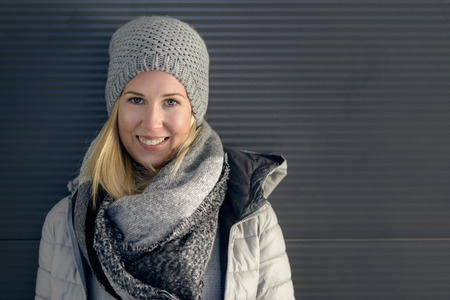Pretty young blond woman in a trendy winter outfit with grey woollen beanie hat, scarf and hooded...