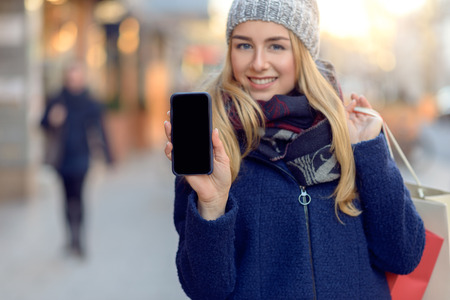 Pretty young woman holding up her mobile phone with a blank display as she stands on an urban street...