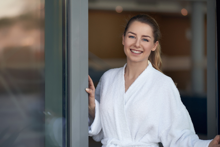 Happy young woman in white bathrobe opening a glass door and smiling to camera, with copy spaceの写真素材