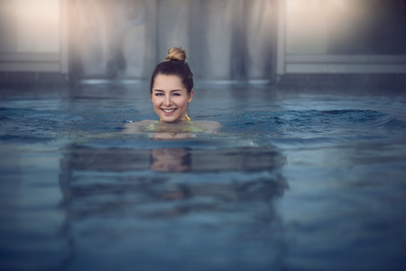 Low angle view of young woman head with hair bun over water surface of indoor swimming pool, smiling...