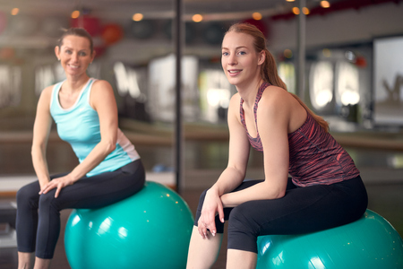 Two healthy young women with pilates or gym balls taking a break from their workout in a gym sitting smiling at the camera in an active lifestyle and fitness conceptの写真素材