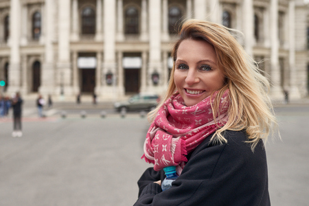 Middle-aged smiling happy woman with blond hair wearing pink scarf and black jacket while standing in front of historic buildingの写真素材