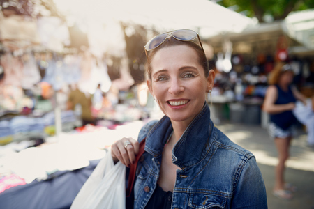 Happy attractive stylish woman shopping at an outdoor market standing looking at the camera with a wide warm friendly smileの写真素材