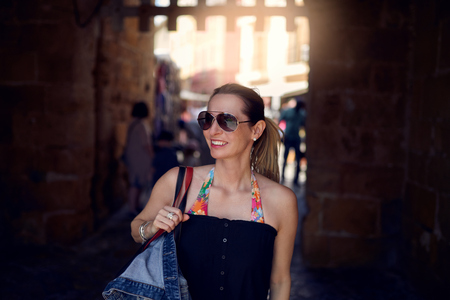 Attractive trendy woman wearing sunglasses and carrying a large handbag over her shoulder shopping in town looking to the side with a smileの写真素材