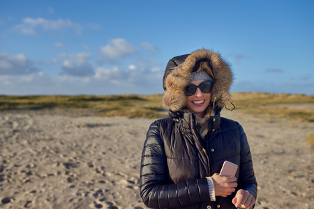 Trendy woman in a warm autumn outfit with sunglasses standing on a beach at sunset casting a long...
