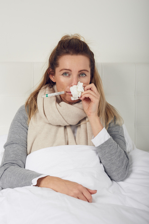 Sick woman with seasonal cold and flu lying wrapped up warmly in bed with a miserable expression blowing her nose on a tissueの写真素材