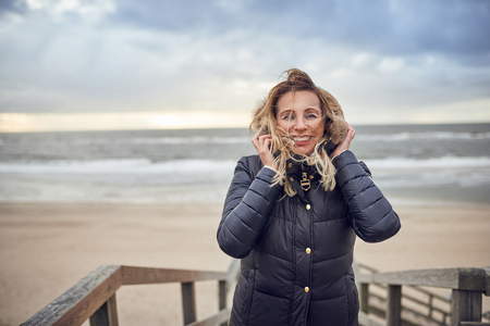 Middle-aged woman braving a cold winter day at the seaside standing on a wooden deck overlooking the...