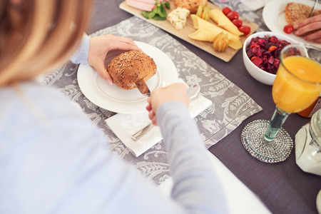 Woman slicing a healthy wholegrain bread roll on a plate at the table with an assortment of cold meats and cheese on a platter in front of herの写真素材