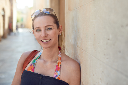 Attractive stylish woman with her sunglasses on her forehead relaxing leaning against a wall in a narrow urban lane smiling at the cameraの写真素材