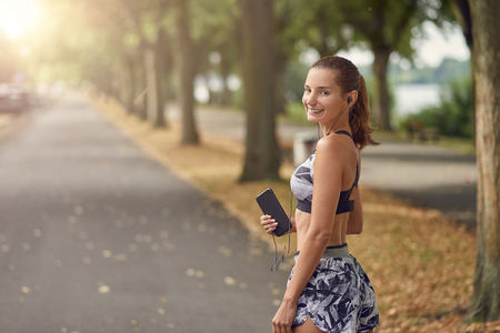 Attractive sporty woman listening to music on her mobile phone out jogging on a tree lined road turning to smile happily at the cameraの写真素材