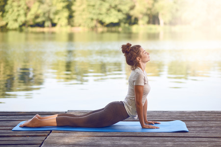 Attractive young woman working out on mat on a wooden deck above a lake or river doing a yoga pose with her eyes closed and a serene expressionの写真素材