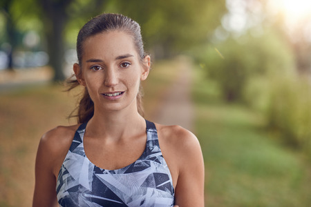 Attractive young woman iin sports attire and a ponytail standing outdoors in a rural park looking pensively at the camera in a close up head and shoulders portraitの写真素材