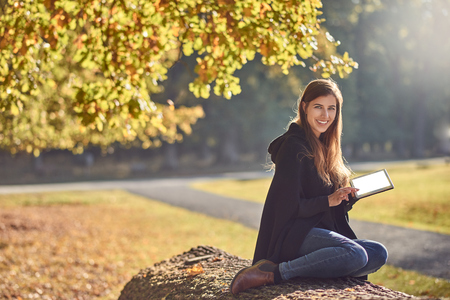 Pretty young woman relaxing in an autumn park reading on her tablet pc and smiling happily at the cameraの写真素材