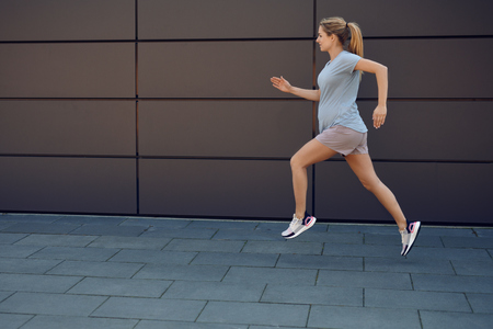 Pregnant young woman keeping fit sprinting along a paved sidewalk in front of an urban wall in a healthy lifestyle conceptの写真素材