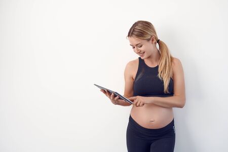 Happy young pregnant woman with bare baby bump or abdomen standing reading on a tablet pc with a...