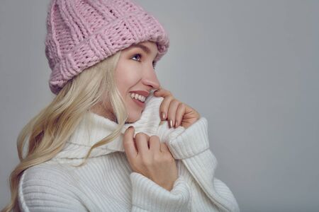 Pretty young blond woman in woollen winter outfit cuddling into the warmth of her polo neck sweater with a happy smile while wearing a pink knitted cap, profile portrait over grey with copy spaceの写真素材