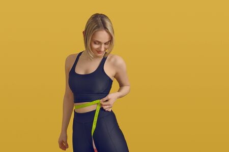 Slim sporty young woman measuring her waist with a tape measure as she looks down with a satisfied smile in a weight loss, healthy diet and exercise concept over a yellow background with copy spaceの写真素材