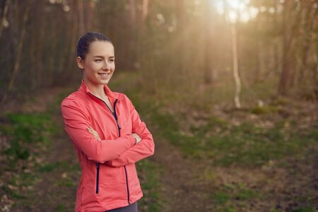 Happy confident young woman with a friendly smile standing with folded arms in woodland backlit by...