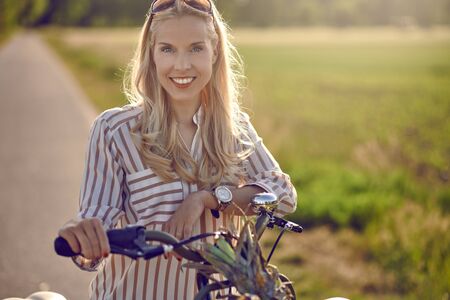 Woman using her bicycle to buy fresh produce standing on a rural road backlit by a warm glow of the sun smiling happily at the camera as she holds her bike with basket full of fruit and vegetablesの写真素材