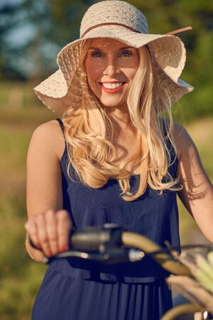 Attractive blond woman with lovely friendly smile standing outdoors with her bicycle in the spring sunshine wearing a broad brimmed straw sunhatの写真素材