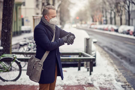 Businessman with shoulder bag wearing a face mask during the Covid-19 pandemic standing on a snowy urban street in winter checking the time on his wristwatchの写真素材