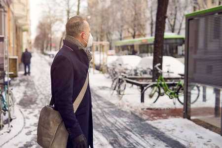 Midde-aged businessman standing at a bus stop, wearing a protective face mask due to corona virus, waiting for his bus to bring him to workの写真素材