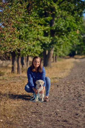 Happy brunette middle-aged woman standing with her little Labrador puppy while kneeing on a rural pathの写真素材