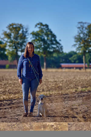 Happy brunette middle-aged woman standing with her little Labrador puppy while kneeing on a rural pathの写真素材