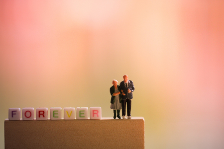 Miniature people: old couple in love standing beside word forever on colorful light background. Happy valentine's day and love conceptの写真素材