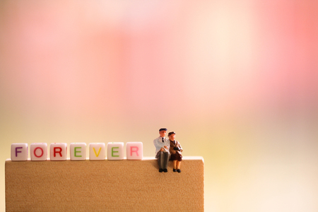 Miniature people: old couple in love sitting beside word forever on colorful light background. Happy valentine's day and love conceptの写真素材