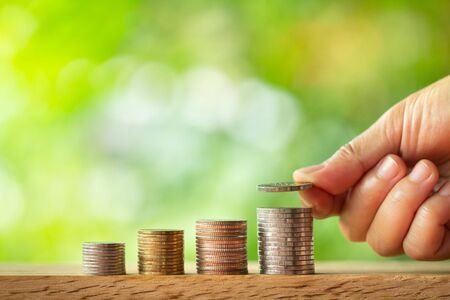 Hand putting coin on coins stack with greenery blurred background. Saving money conceptの写真素材