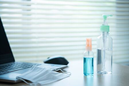 Alcohol spray bottle and sanitary hand cleansing gel on table with surgical mask and laptop computer in office. Virus protection concept.の写真素材