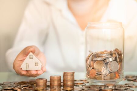 Closeup woman holding house figure with coins stack and glass jar on table. Business and savings money concept.の写真素材